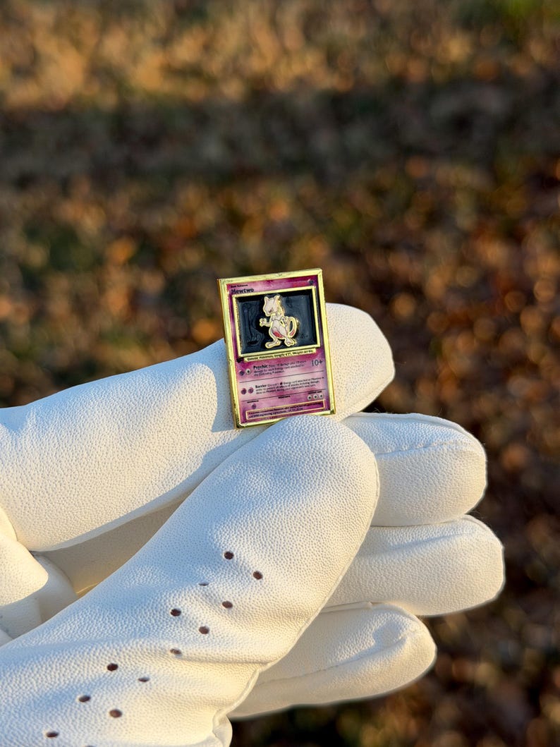 May include: A gold-framed pin featuring a Pokemon card design with a purple and pink Mewtwo illustration. The pin is held by a person wearing a white glove. The background is blurred with natural tones.