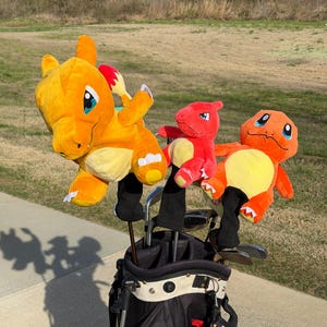 Puede incluir: Una bolsa de golf negra y roja con tres cubiertas de cabeza de palo de golf Charmander de felpa. Las cubiertas de Charmander son naranjas y rojas con vientres amarillos y cubiertas de palo negras. La bolsa de golf está sobre una superficie pavimentada con un fondo de hierba.