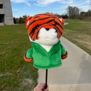 Puede incluir: Una funda para la cabeza del palo de golf con forma de tigre. La funda presenta rayas naranjas y negras, pelaje blanco y una chaqueta verde con ribetes naranjas. Est&aacute; unida a un eje marr&oacute;n oscuro.