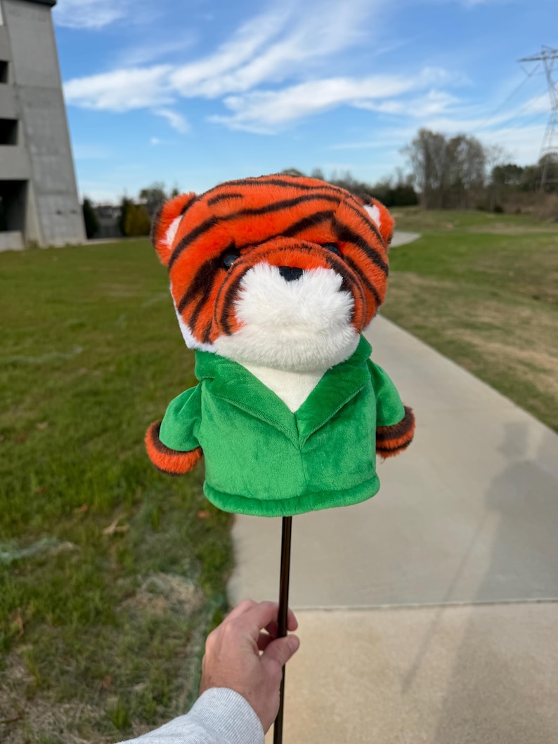 Puede incluir: Una funda para la cabeza del palo de golf con forma de tigre. La funda presenta rayas naranjas y negras, pelaje blanco y una chaqueta verde con ribetes naranjas. Est&aacute; unida a un eje marr&oacute;n oscuro.