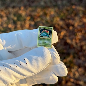 May include: A gold-colored, rectangular trading card featuring a green and blue cartoon character. The card has a green border and text, including the name "Venusaur". The card is held in a white glove.