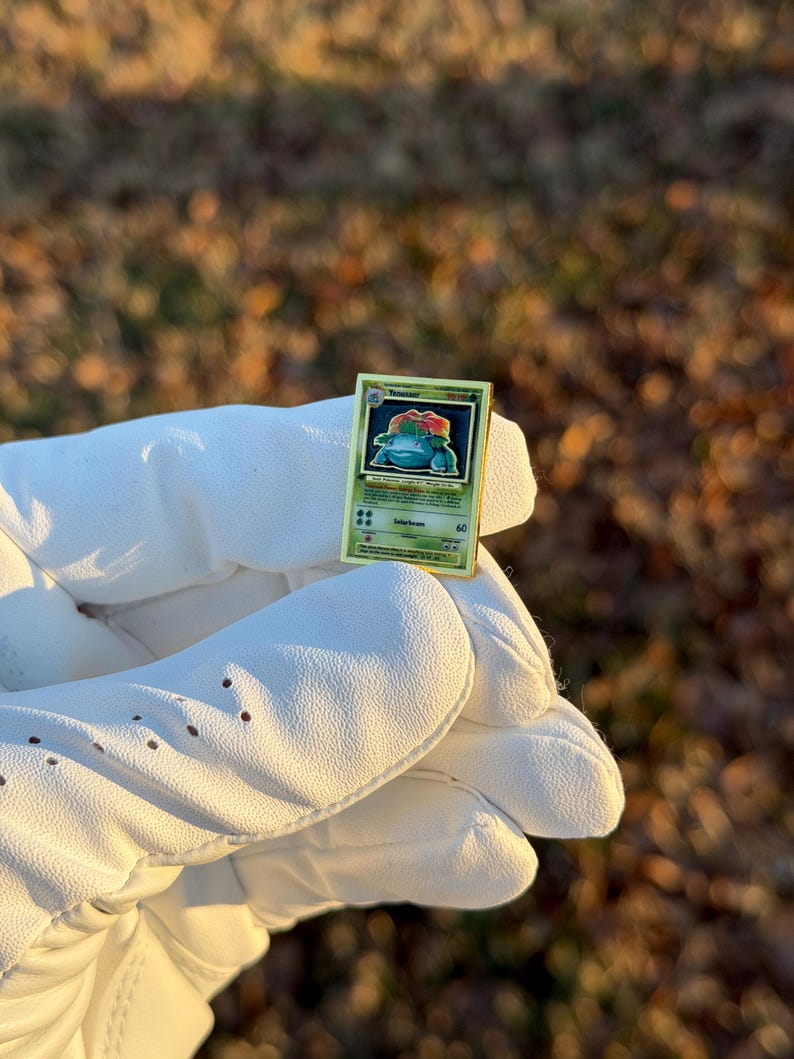 May include: A gold-colored, rectangular trading card featuring a green and blue cartoon character. The card has a green border and text, including the name "Venusaur". The card is held in a white glove.