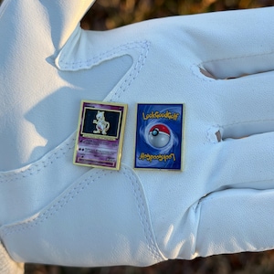 May include: Two gold-framed Pokemon card pins. One features a purple Mewtwo character, the other has a blue background with a Poke Ball and the text "LookGoodGolf". The pins are displayed on a white surface.