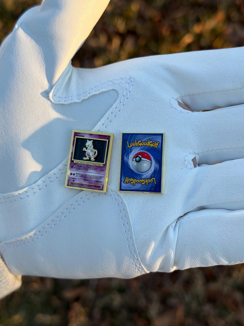 May include: Two gold-framed Pokemon card pins. One features a purple Mewtwo character, the other has a blue background with a Poke Ball and the text "LookGoodGolf". The pins are displayed on a white surface.
