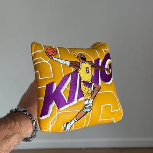 Puede incluir: Funda para putter amarilla y morada con un jugador de baloncesto haciendo un mate. La palabra "KING" está bordada en morado, con el número 6 en la camiseta del jugador. La funda tiene un diseño geométrico.