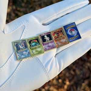 May include: Five Pokemon trading card pins with gold borders are displayed. The pins feature various Pokemon characters and the text "LookGoodGolf". The pins are arranged on a white surface, with a blurred background of green and brown.