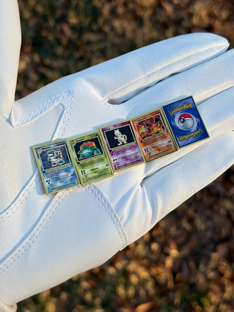 May include: Five Pokemon trading card pins with gold borders are displayed. The pins feature various Pokemon characters and the text "LookGoodGolf". The pins are arranged on a white surface, with a blurred background of green and brown.