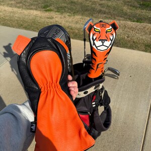 May include: An orange and black golf club head cover with a tiger face design. The cover is held next to a golf bag with other clubs. A matching orange and black golf glove is also visible.