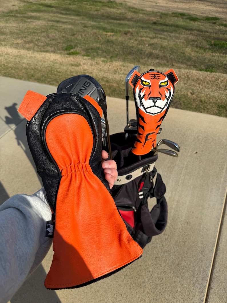 May include: An orange and black golf club head cover with a tiger face design. The cover is held next to a golf bag with other clubs. A matching orange and black golf glove is also visible.