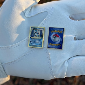 May include: Two gold-framed pins. One pin features a blue and gold Pokemon card design with a Blastoise illustration. The other pin has a blue background with a Poke Ball and the text "LookGoodGolf".