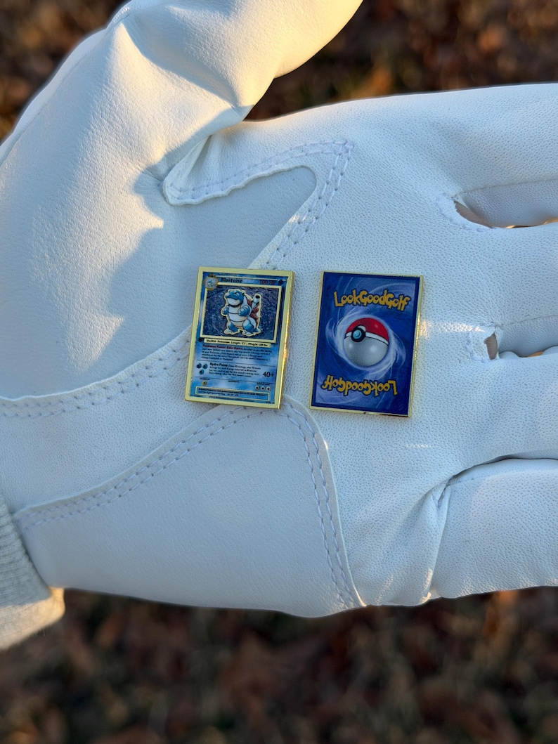 May include: Two gold-framed pins. One pin features a blue and gold Pokemon card design with a Blastoise illustration. The other pin has a blue background with a Poke Ball and the text "LookGoodGolf".
