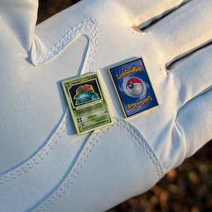 May include: Two small, rectangular pins featuring Pokemon card designs. One pin shows a green and yellow card with a cartoon plant creature. The other pin has a blue background with a Poke Ball and the text "Look Good Golf".