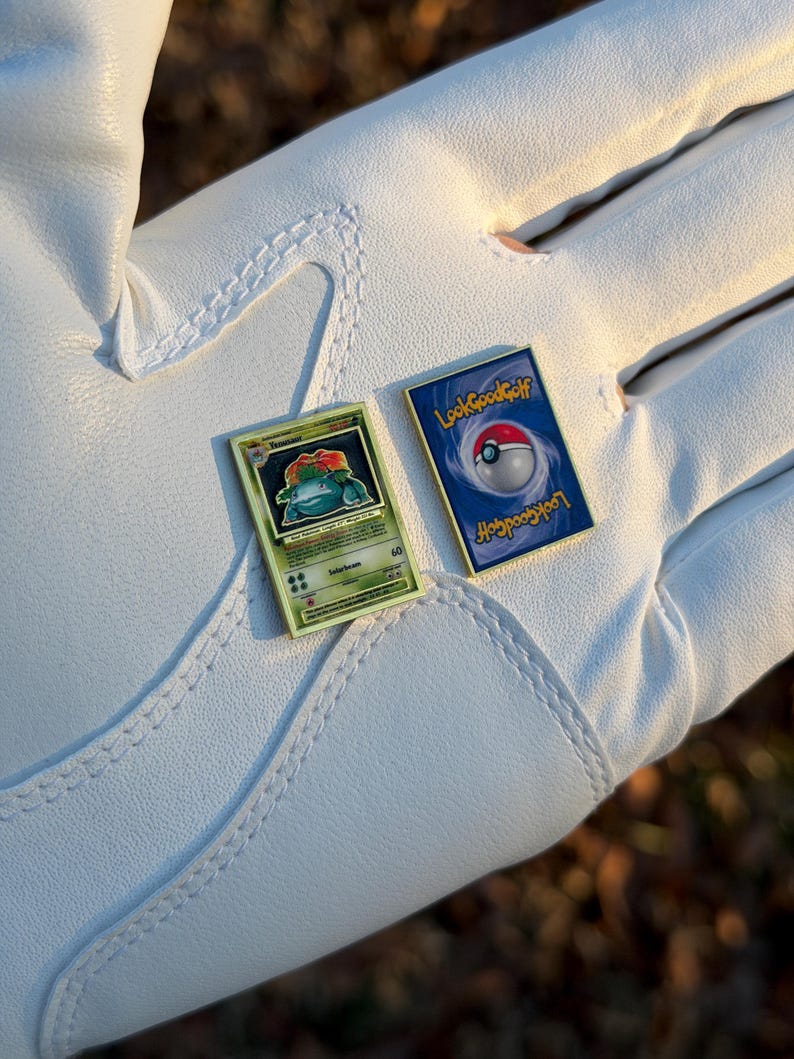 May include: Two small, rectangular pins featuring Pokemon card designs. One pin shows a green and yellow card with a cartoon plant creature. The other pin has a blue background with a Poke Ball and the text "Look Good Golf".