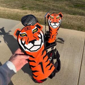 May include: Golf club head covers with a tiger design. The covers are orange with black stripes and a white face. The golf bag is black and red. The PING club is visible.