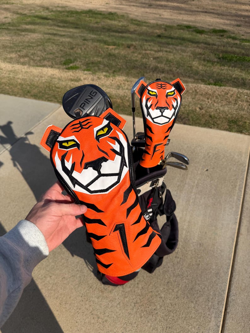 May include: Golf club head covers with a tiger design. The covers are orange with black stripes and a white face. The golf bag is black and red. The PING club is visible.