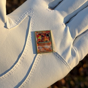 May include: A gold-framed Charizard Pok&eacute;mon trading card. The card features a fiery orange and red illustration of Charizard. The card is held in a white golf glove.
