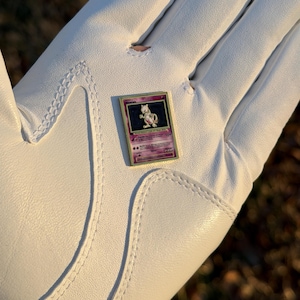 May include: A Pokemon trading card featuring Mewtwo, with a purple and gold design. The card is held in a white golf glove. The card has a purple background with the name "Mewtwo" at the top.