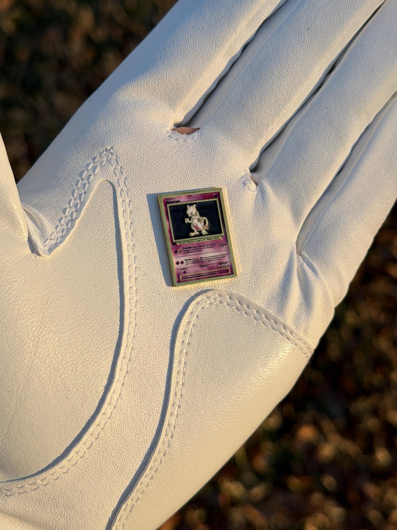 May include: A Pokemon trading card featuring Mewtwo, with a purple and gold design. The card is held in a white golf glove. The card has a purple background with the name "Mewtwo" at the top.