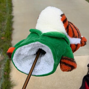 Puede incluir: Una funda para la cabeza del palo de golf con forma de tigre. La funda tiene una chaqueta verde, piel blanca y rayas naranjas y negras. El palo de golf marr&oacute;n est&aacute; insertado en la funda.