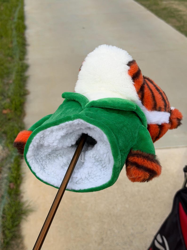 Puede incluir: Una funda para la cabeza del palo de golf con forma de tigre. La funda tiene una chaqueta verde, piel blanca y rayas naranjas y negras. El palo de golf marr&oacute;n est&aacute; insertado en la funda.