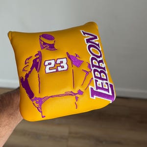 May include: Yellow and purple golf club head cover. The cover features a purple embroidered image of a basketball player with the number 23 and the name LEBRON. The cover is made of a smooth, leather-like material.