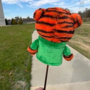 Puede incluir: Una funda para palo de golf con cabeza de tigre de peluche con rayas naranjas y negras, chaqueta verde y eje marr&oacute;n. La funda se sostiene sobre un fondo de cielo azul, hierba verde y un camino de hormig&oacute;n.