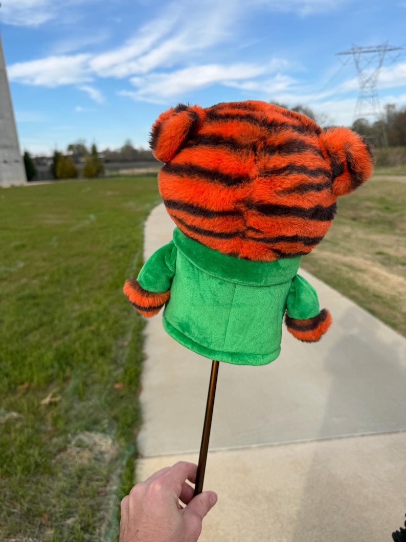 Puede incluir: Una funda para palo de golf con cabeza de tigre de peluche con rayas naranjas y negras, chaqueta verde y eje marr&oacute;n. La funda se sostiene sobre un fondo de cielo azul, hierba verde y un camino de hormig&oacute;n.