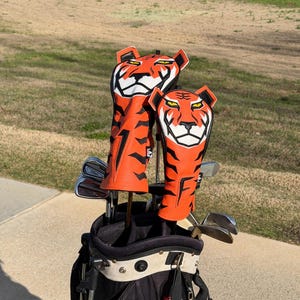 May include: A golf bag with clubs and two orange and black tiger-themed head covers. The head covers have a tiger face design with white and black accents. The golf bag is black and red.