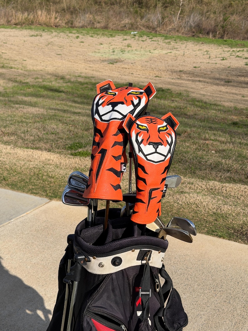 May include: A golf bag with clubs and two orange and black tiger-themed head covers. The head covers have a tiger face design with white and black accents. The golf bag is black and red.