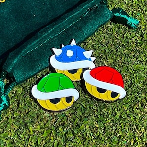 May include: Three colorful enamel pins shaped like the iconic turtle shells from the Super Mario Bros. video game. The pins are blue, green, and red.