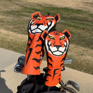 May include: Two orange and black tiger-themed golf club head covers. The covers are made of leather and feature a tiger face design with white and black accents. The covers are in a golf bag with golf clubs.