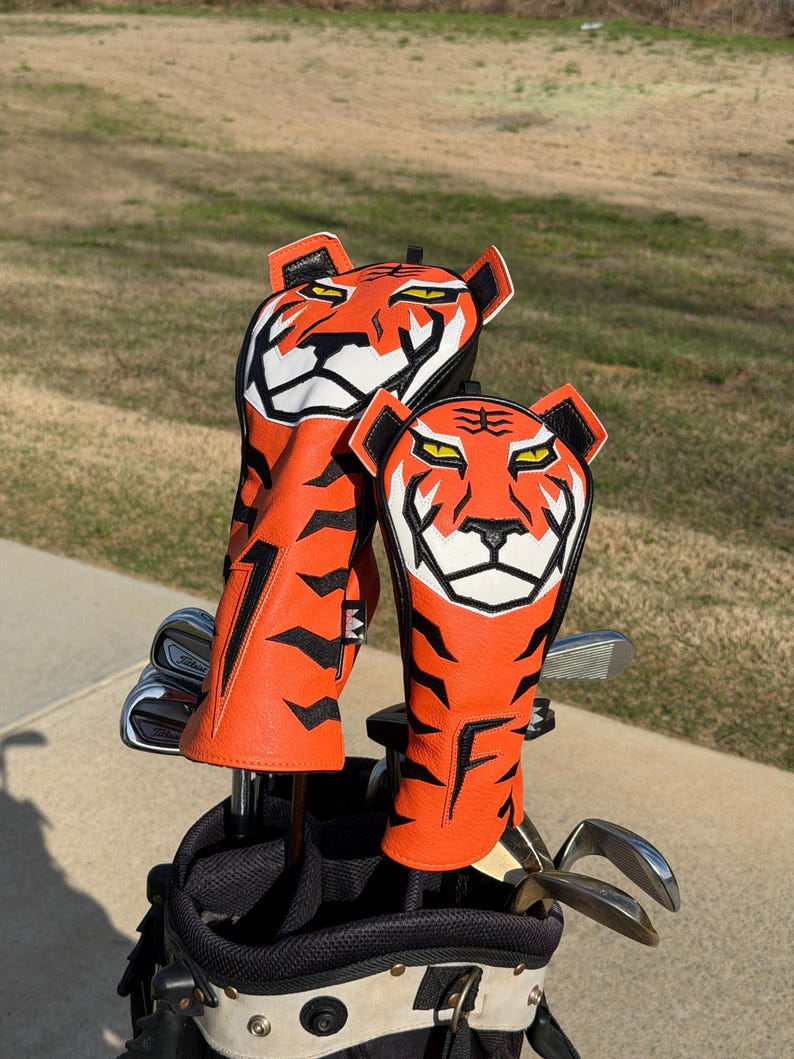 May include: Two orange and black tiger-themed golf club head covers. The covers are made of leather and feature a tiger face design with white and black accents. The covers are in a golf bag with golf clubs.