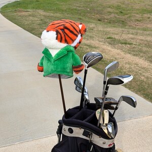Puede incluir: Una bolsa de golf con una funda para la cabeza del palo con tem&aacute;tica de tigre. La funda es de rayas naranjas y negras con una chaqueta verde. La bolsa contiene varios palos de golf, colocados en un camino pavimentado con un campo de golf al fondo.