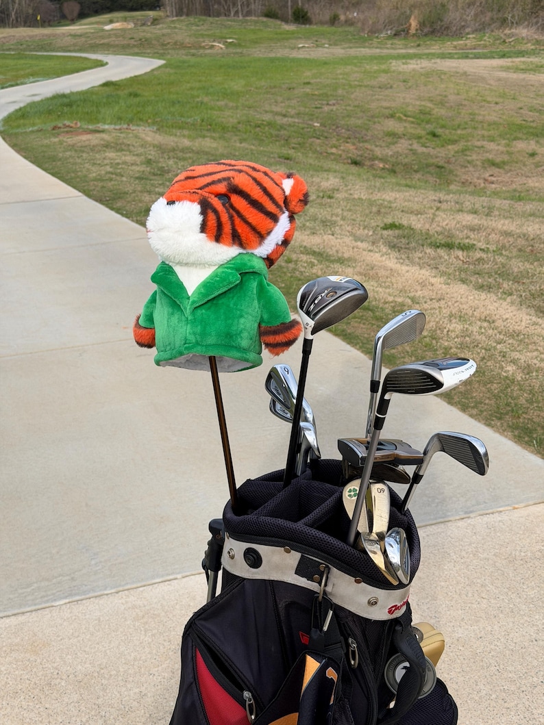 Puede incluir: Una bolsa de golf con una funda para la cabeza del palo con tem&aacute;tica de tigre. La funda es de rayas naranjas y negras con una chaqueta verde. La bolsa contiene varios palos de golf, colocados en un camino pavimentado con un campo de golf al fondo.