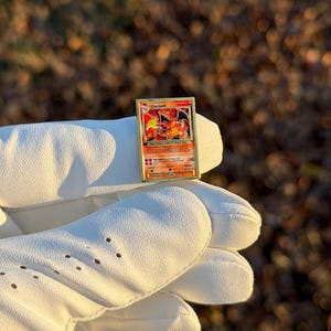 May include: A Charizard Pok&eacute;mon trading card with a red, orange, and yellow illustration of a dragon-like creature. The card is held by a white glove. The card has the number 100 in the bottom right corner.