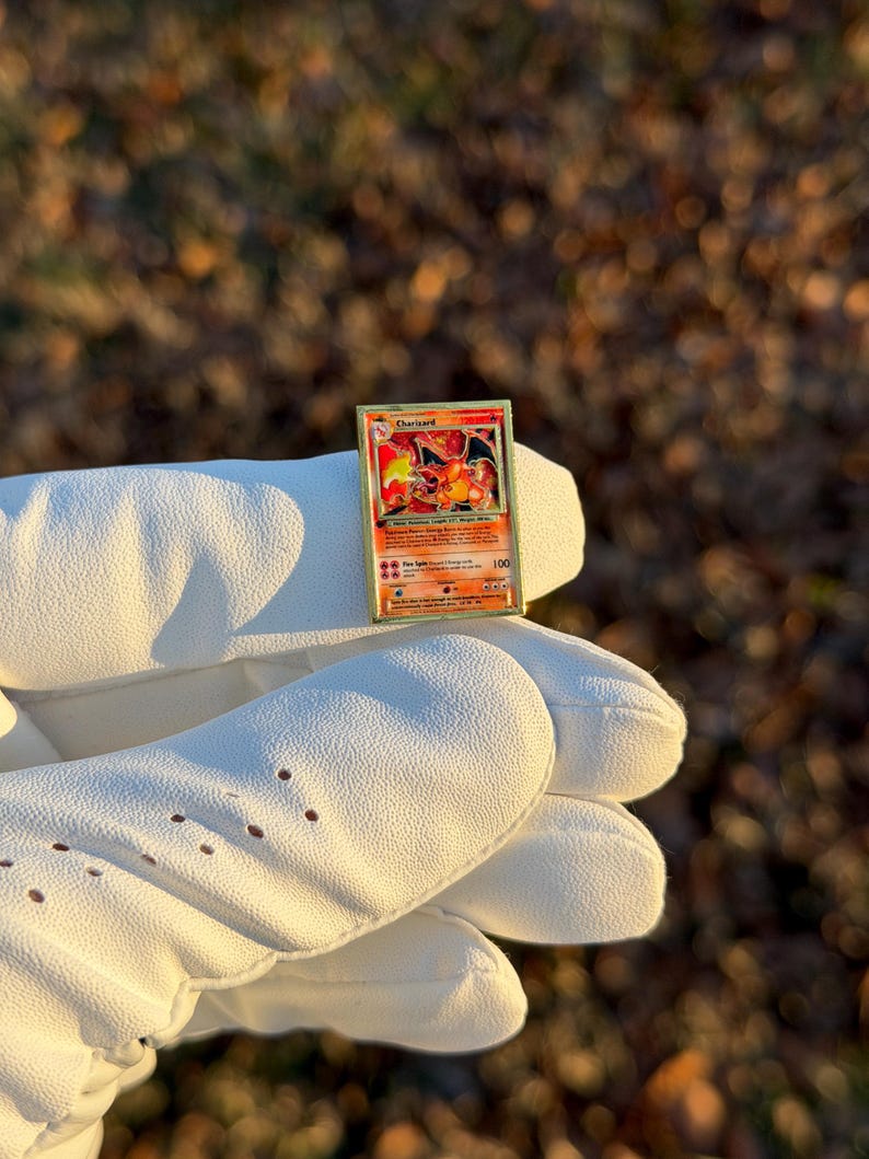 May include: A Charizard Pok&eacute;mon trading card with a red, orange, and yellow illustration of a dragon-like creature. The card is held by a white glove. The card has the number 100 in the bottom right corner.