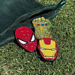 May include: Three enamel pins featuring popular superhero characters. The pins are gold, red, and yellow. The gold pin is a hand with gems on the fingers. The red pin is a red and black Spiderman mask. The yellow pin is a yellow and red Iron Man mask.