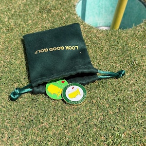 May include: A dark green velvet pouch with gold text reading "LOOK GOOD GOLF". Two golf ball markers are visible, one green and yellow with a map design and the other white and green with "Masters Tournament" text. The items are on green grass.