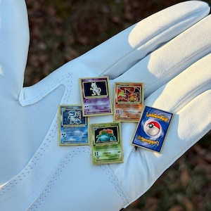 May include: A collection of Pokemon trading card pins, including Charizard, Blastoise, Venusaur, and Mewtwo, with a LookGoodGolf pin. The pins are gold-framed and displayed on a white glove.