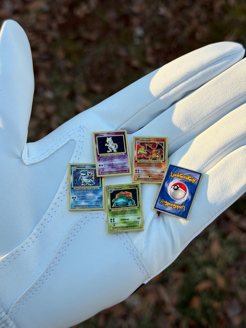 May include: A collection of Pokemon trading card pins, including Charizard, Blastoise, Venusaur, and Mewtwo, with a LookGoodGolf pin. The pins are gold-framed and displayed on a white glove.