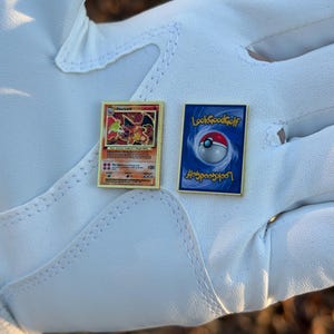 May include: Two gold-framed pins featuring a Charizard Pokemon card and a Poke Ball design with the text "LookGoodGolf" on a white surface. The Charizard pin has a red and orange dragon illustration.