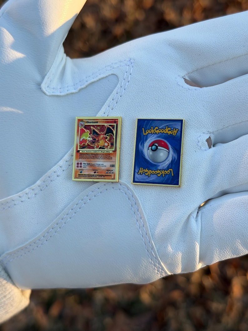May include: Two gold-framed pins featuring a Charizard Pokemon card and a Poke Ball design with the text "LookGoodGolf" on a white surface. The Charizard pin has a red and orange dragon illustration.