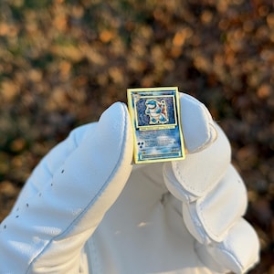 May include: A gold-bordered Pok&eacute;mon trading card featuring the character Blastoise. The card has a blue and white illustration of Blastoise, with text at the bottom. The card is held in a white golf glove against a blurred background of fall foliage.