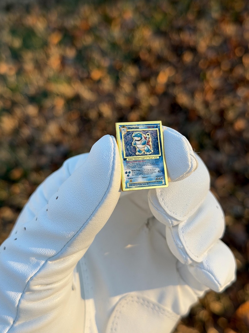 May include: A gold-bordered Pok&eacute;mon trading card featuring the character Blastoise. The card has a blue and white illustration of Blastoise, with text at the bottom. The card is held in a white golf glove against a blurred background of fall foliage.