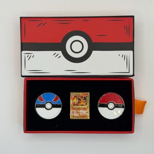 May include: A red and white box resembling a Pok&eacute; Ball, open to reveal a gold Charizard trading card and two Pok&eacute; Ball pins. The pins are red and white, and blue and white. The box has a black interior.