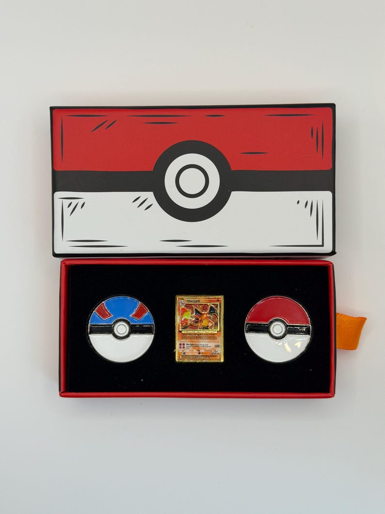 May include: A red and white box resembling a Pok&eacute; Ball, open to reveal a gold Charizard trading card and two Pok&eacute; Ball pins. The pins are red and white, and blue and white. The box has a black interior.