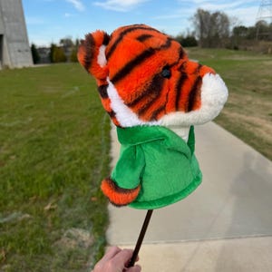 Puede incluir: Una funda para palo de golf con forma de cabeza de tigre, con rayas naranjas y negras, detalles blancos y una chaqueta verde. La funda se sostiene sobre un fondo de hierba verde y cielo azul.