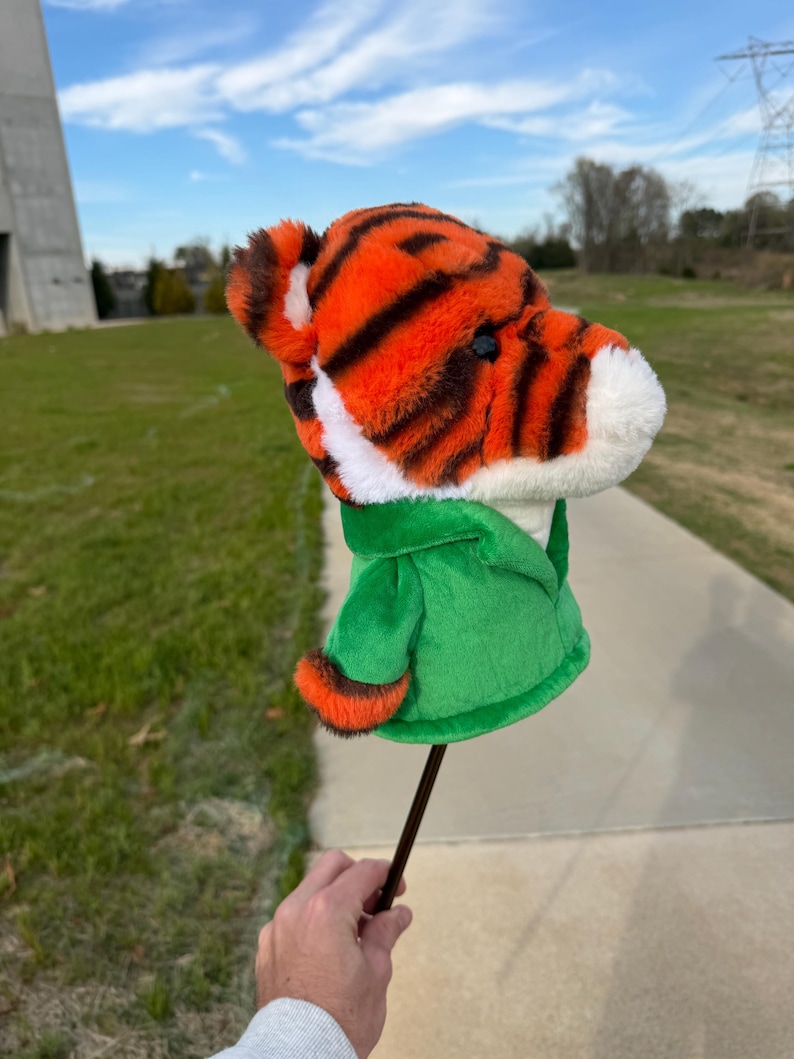 Puede incluir: Una funda para palo de golf con forma de cabeza de tigre, con rayas naranjas y negras, detalles blancos y una chaqueta verde. La funda se sostiene sobre un fondo de hierba verde y cielo azul.