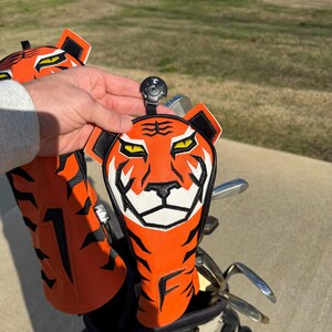 May include: Orange and black tiger-faced golf club head covers. The covers have a white and black face design, with the letter "F" on the bottom. The golf club head covers are in a golf bag with golf clubs.