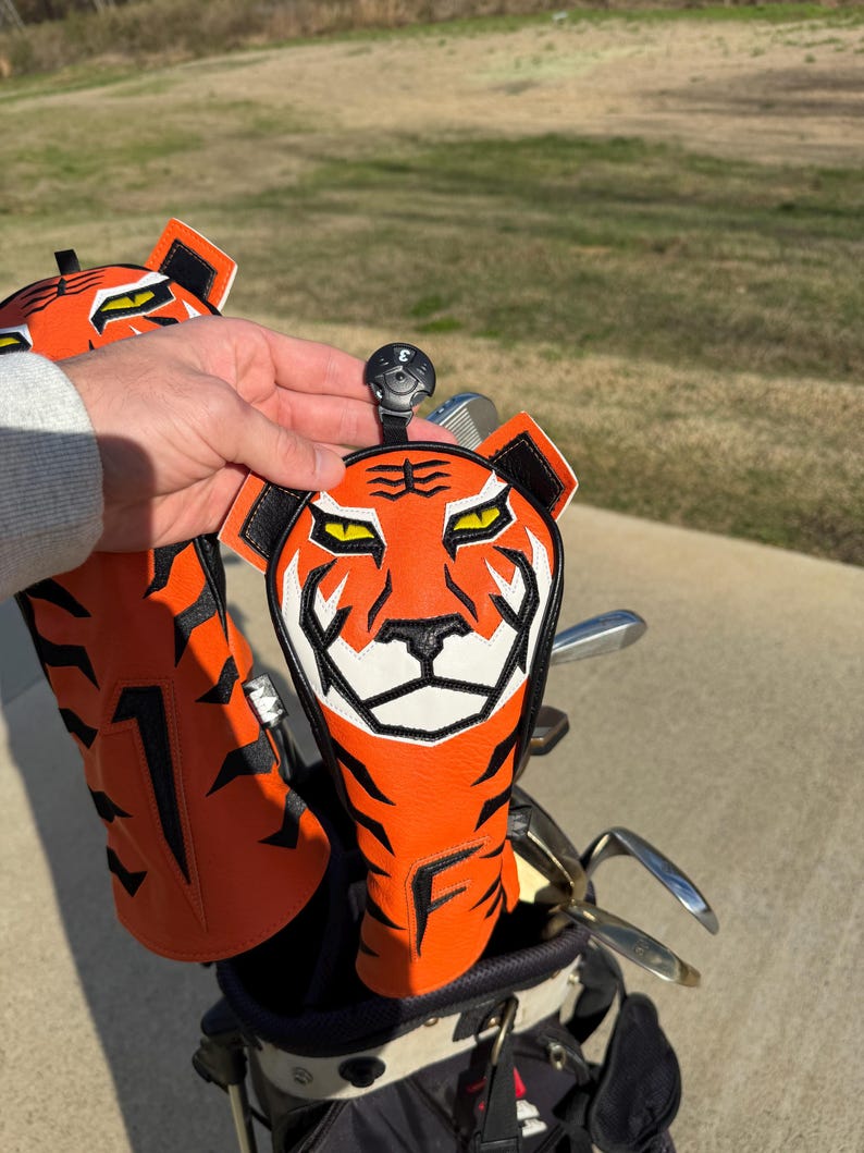 May include: Orange and black tiger-faced golf club head covers. The covers have a white and black face design, with the letter "F" on the bottom. The golf club head covers are in a golf bag with golf clubs.